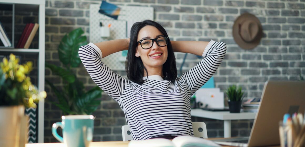 Relaxed woman leaning back at her desk with hands behind her head, symbolising calm productivity.
